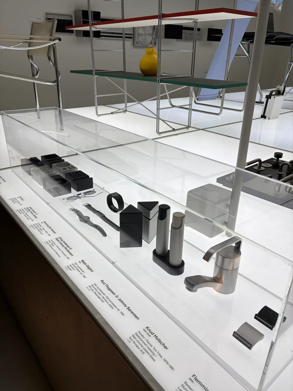A glass museum case of small cast-metal objects, with a red-and-green steel shelving unit and a tubular chair behind it.