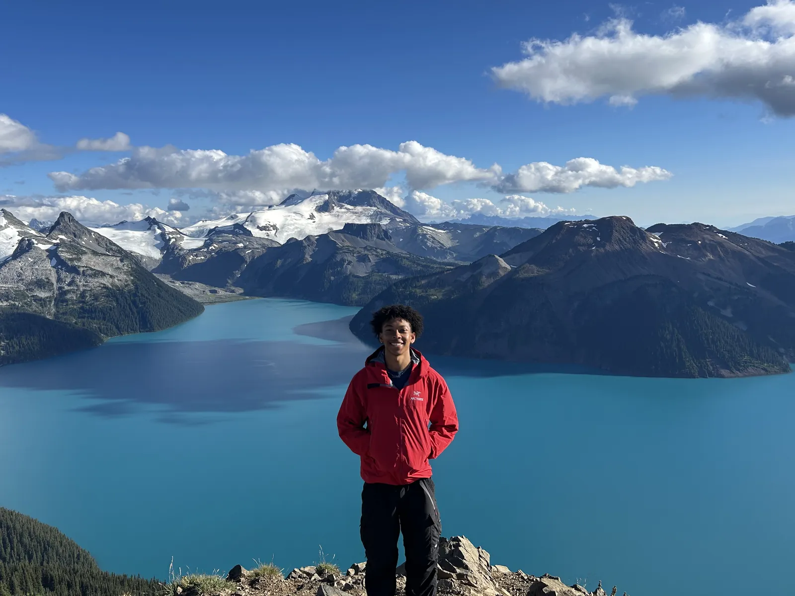 Evan in a red jacket on a rocky summit above a turquoise alpine lake, with snow-capped peaks behind.