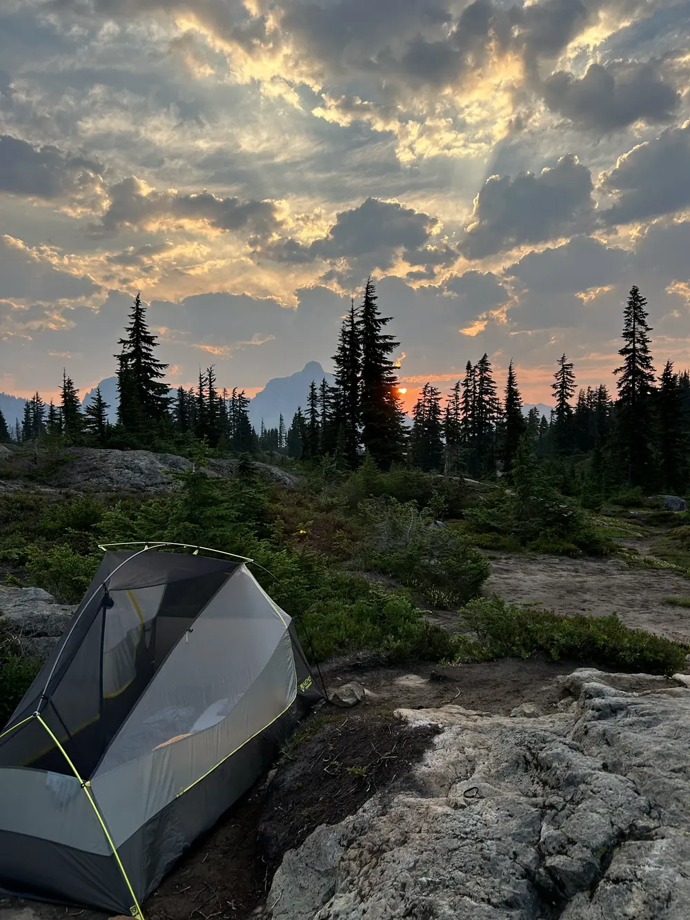 A small backpacking tent on bare rock at sunrise, with subalpine firs silhouetted against an orange sky.
